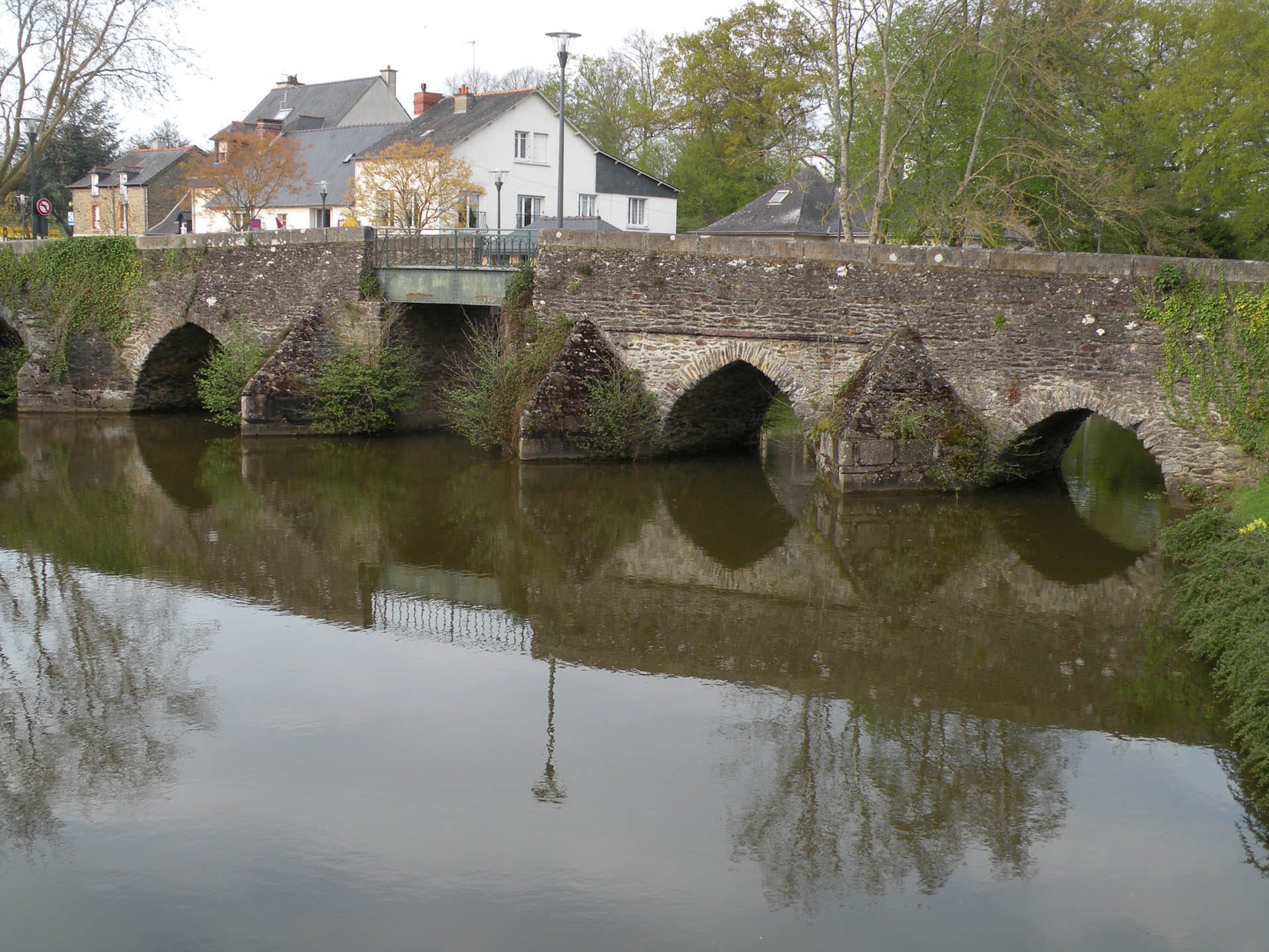 Old stone arch bridge with five arches reflected in calm river water, with houses and trees visible behind.